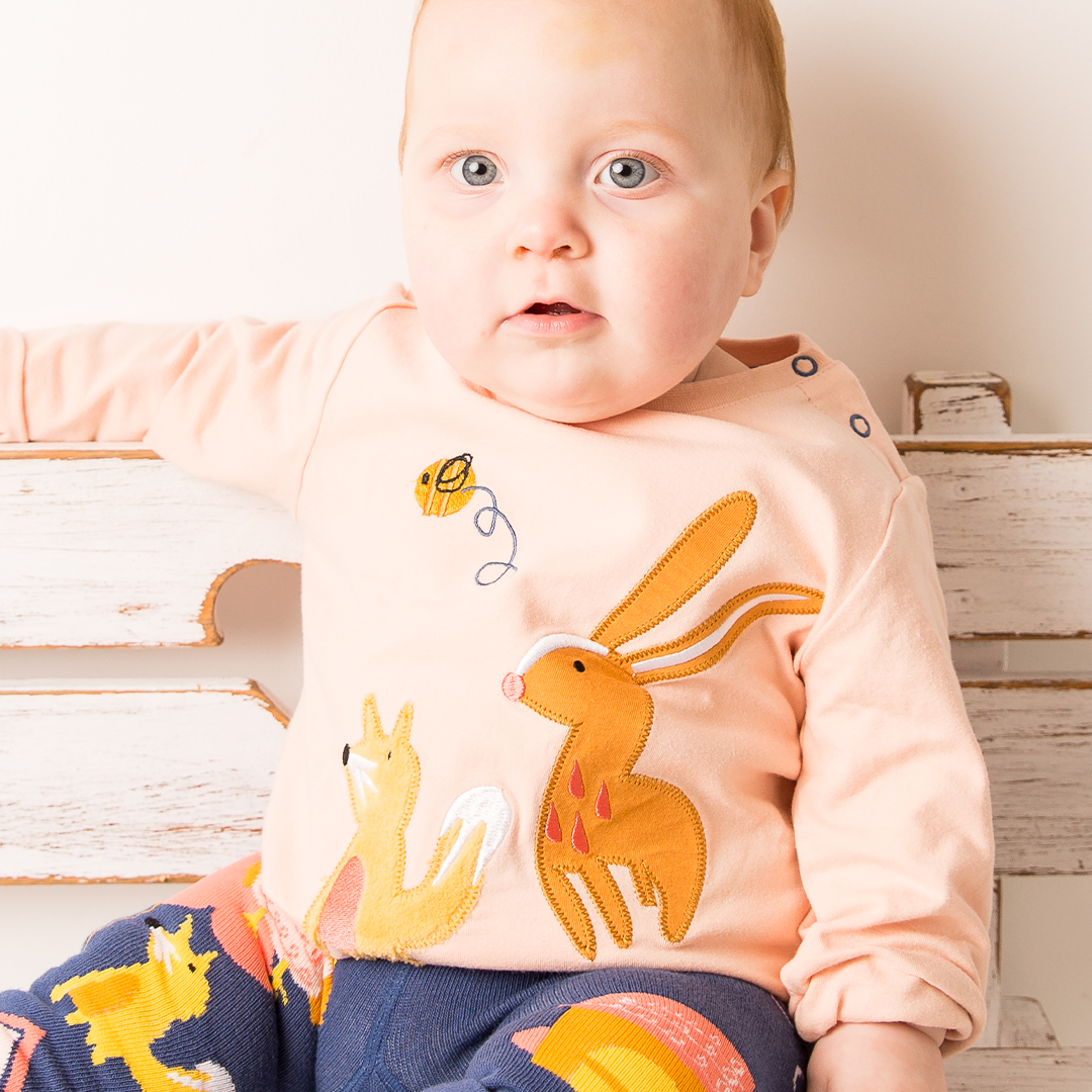 Baby wearing a light pink long-sleeve shirt with animal prints and dark blue leggings with colorful patterns, sitting on a wooden floor.